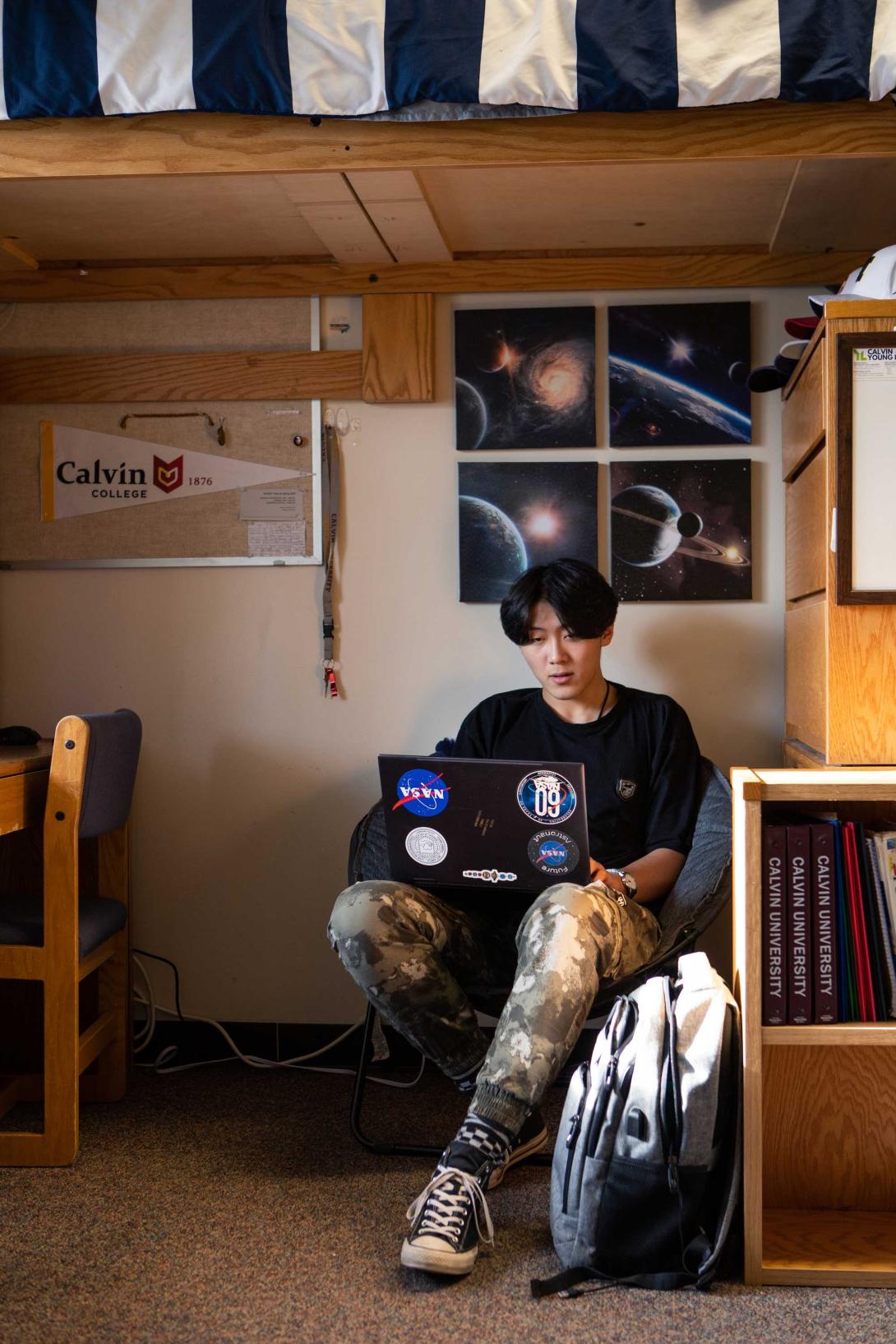 A student works on his computer at his dorm room desk, with pictures of outer space on the wall, a calvin university penant on the bulletin board, and his backpack on the ground.