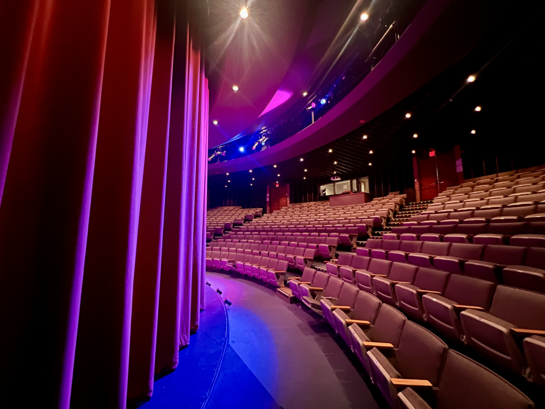 A view of the Gezon auditorium, photographed from stage right. The curtains are down and lit in blues and purples.