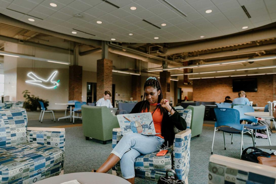 A student works on her laptop in a chair in the Devos communication building lobby.