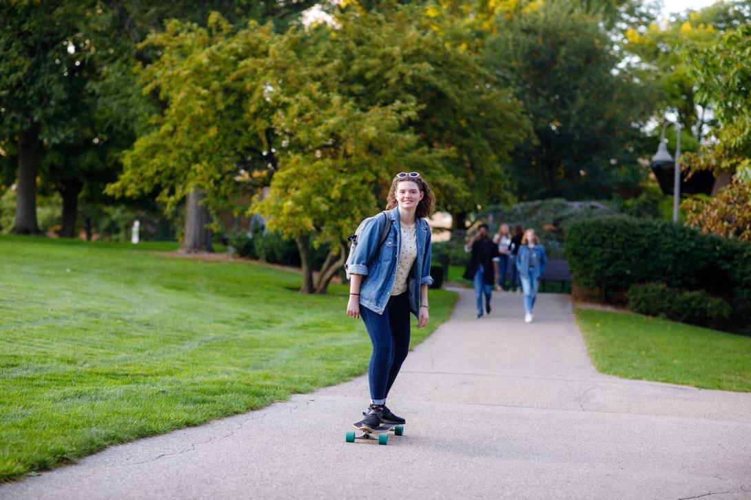 A student long boards down a paved path through a green lawn on Calvin University's campus.
