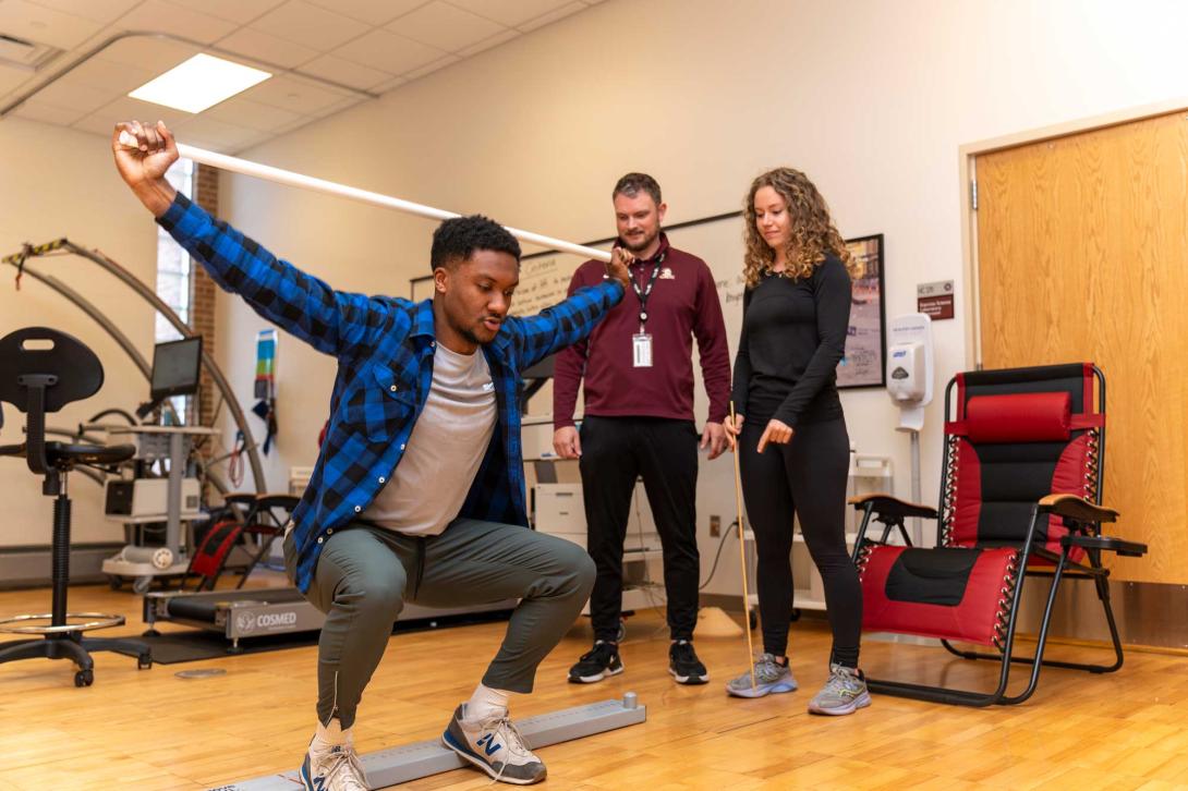 A kinesiology student squats while stretching a band with his arms raised, while another student and professor observe.