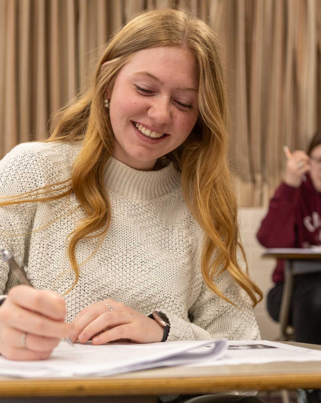 A student writes in a notebook and smiles at her desk, with a classmate at her desk in the background.