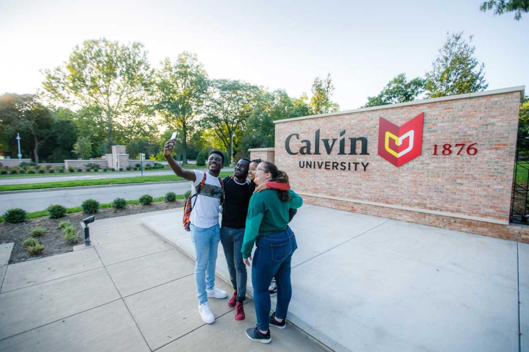 Four students take a selfie in front of the Calvin University brick entrance sign.