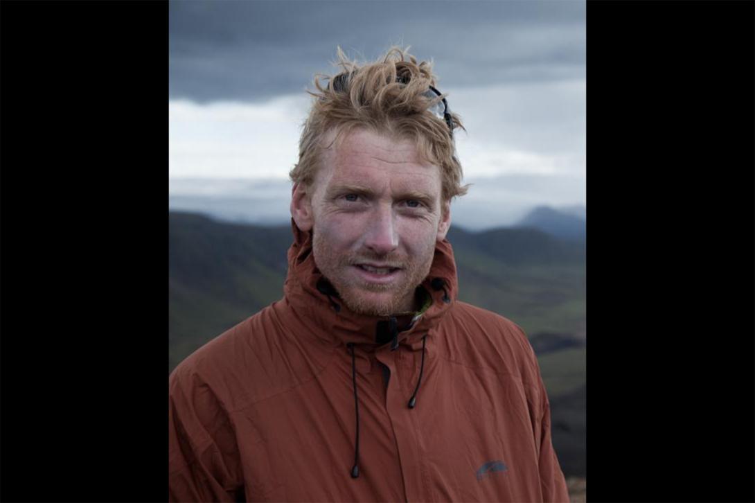 Alastair Humphreys poses in an orange jacket under a cloudy sky with mountains in the background.