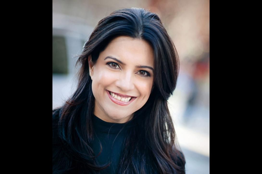 Reshma, an Indian American woman with long, straight black hair smiles for a professional headshot wearing a black shirt with a blurred outdoor scene in the background.