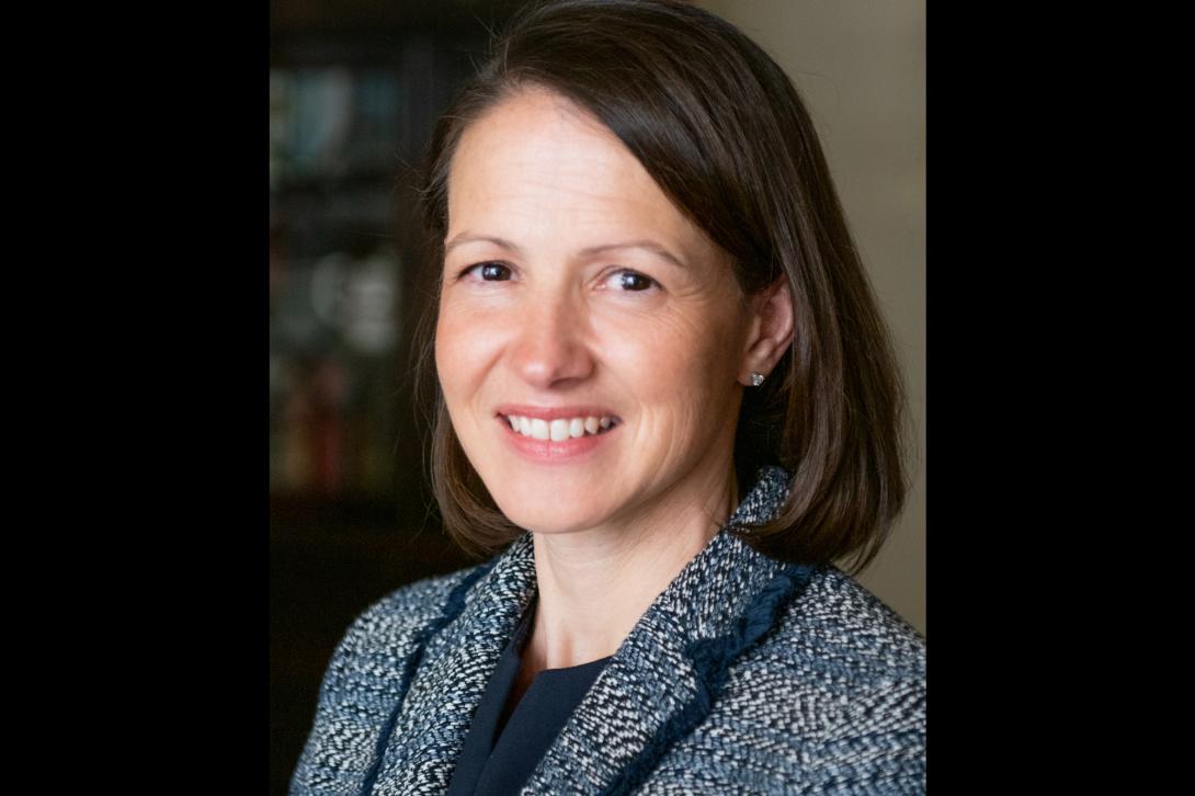 Heather, a white woman with shoulder-length, straight brown hair, smiles for a professional headshot wearing a black and white suit jacket.