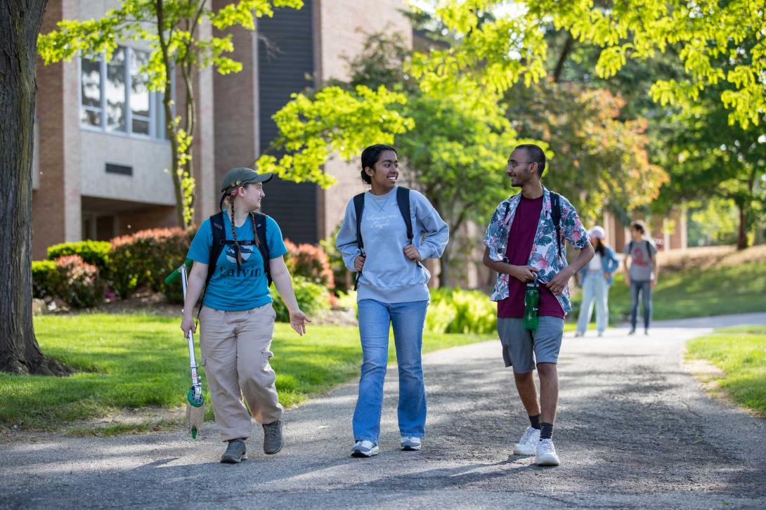 Three students walk down the path chatting and smiling at Calvin University, one holds a scooter.