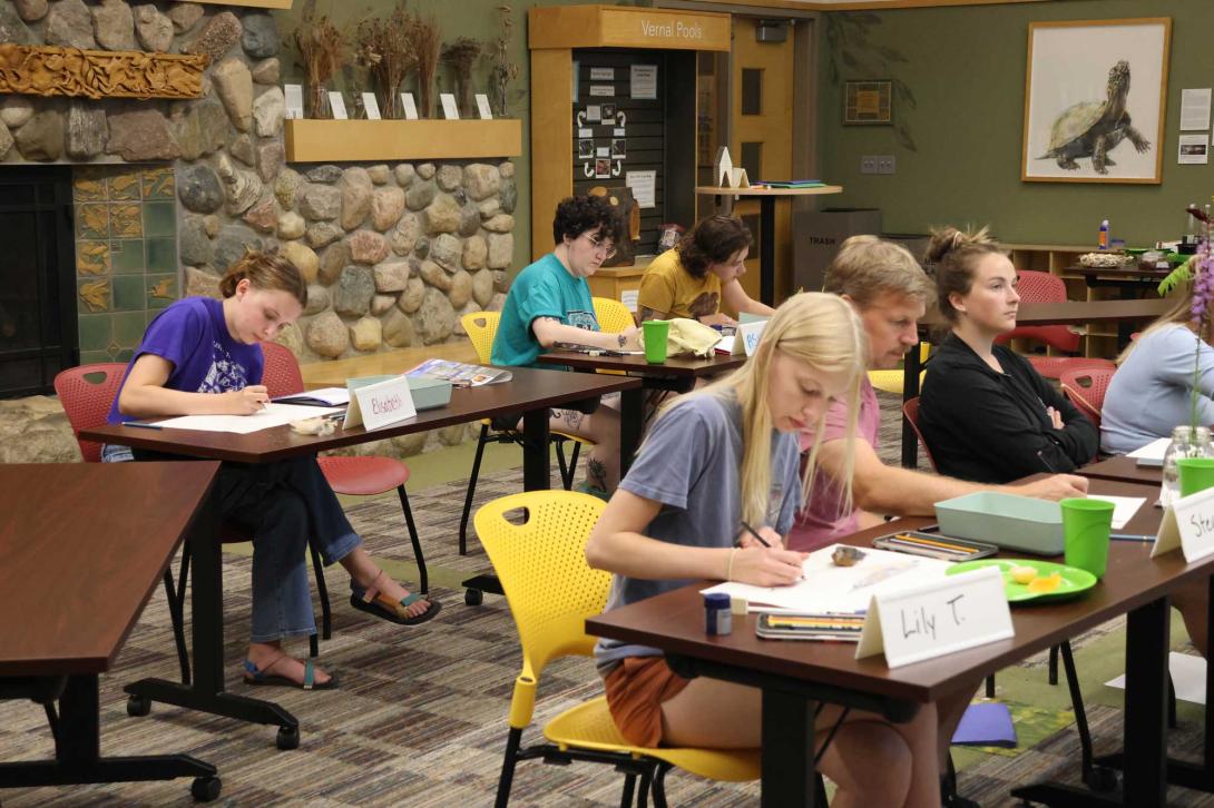Students write at tables with a fireplace behind them.