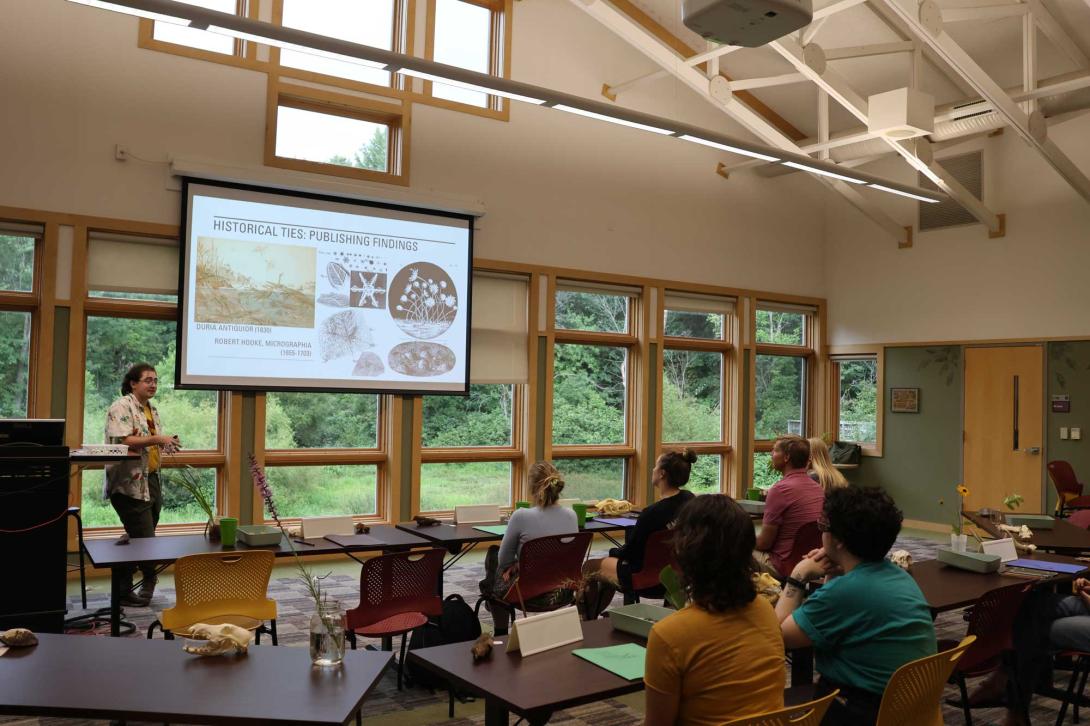 Someone leads a lecture for students in a room with high vaulted ceilings and a wall of windows looking out over green woods.