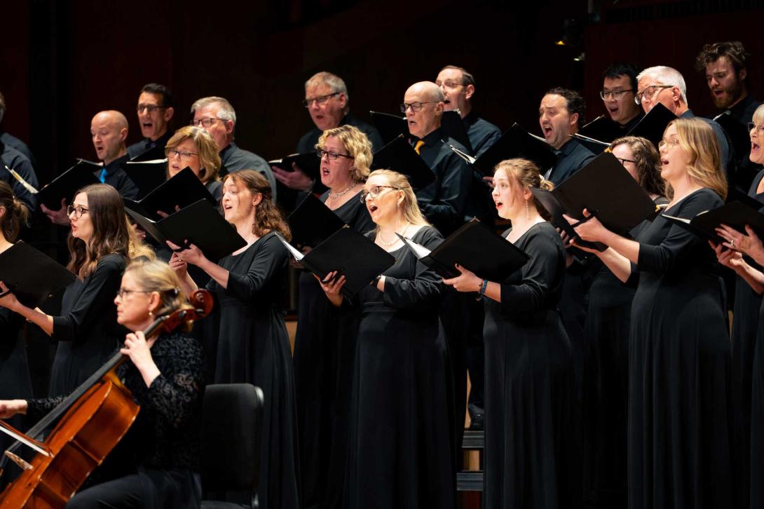 A choir performs with a cello on stage at Calvin University, dressed in black dresses and tuxedos, holding folders.