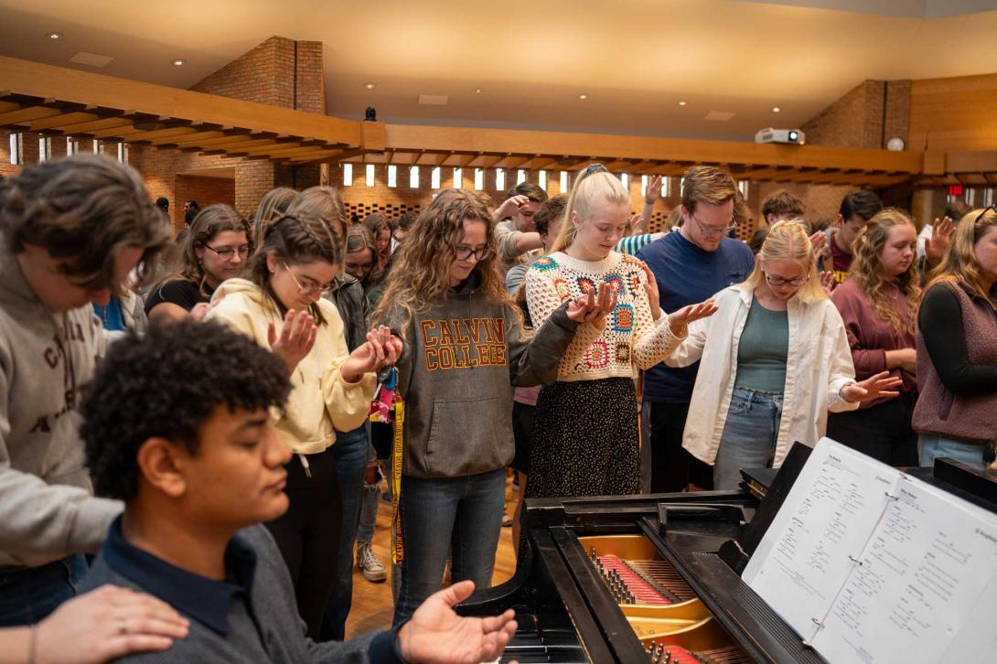 Students pray with hands open, gathered around the piano in the Calvin chapel.