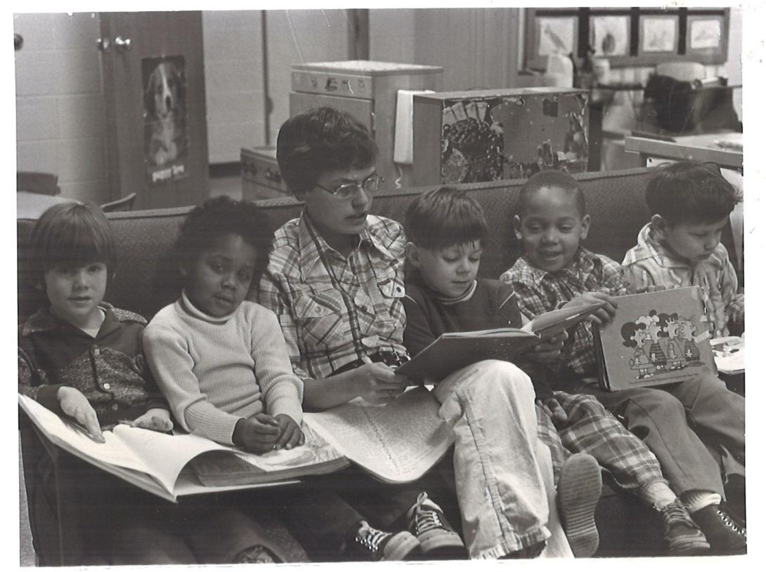 Student reading to five school-aged children on a couch, vintage photo from the 80s
