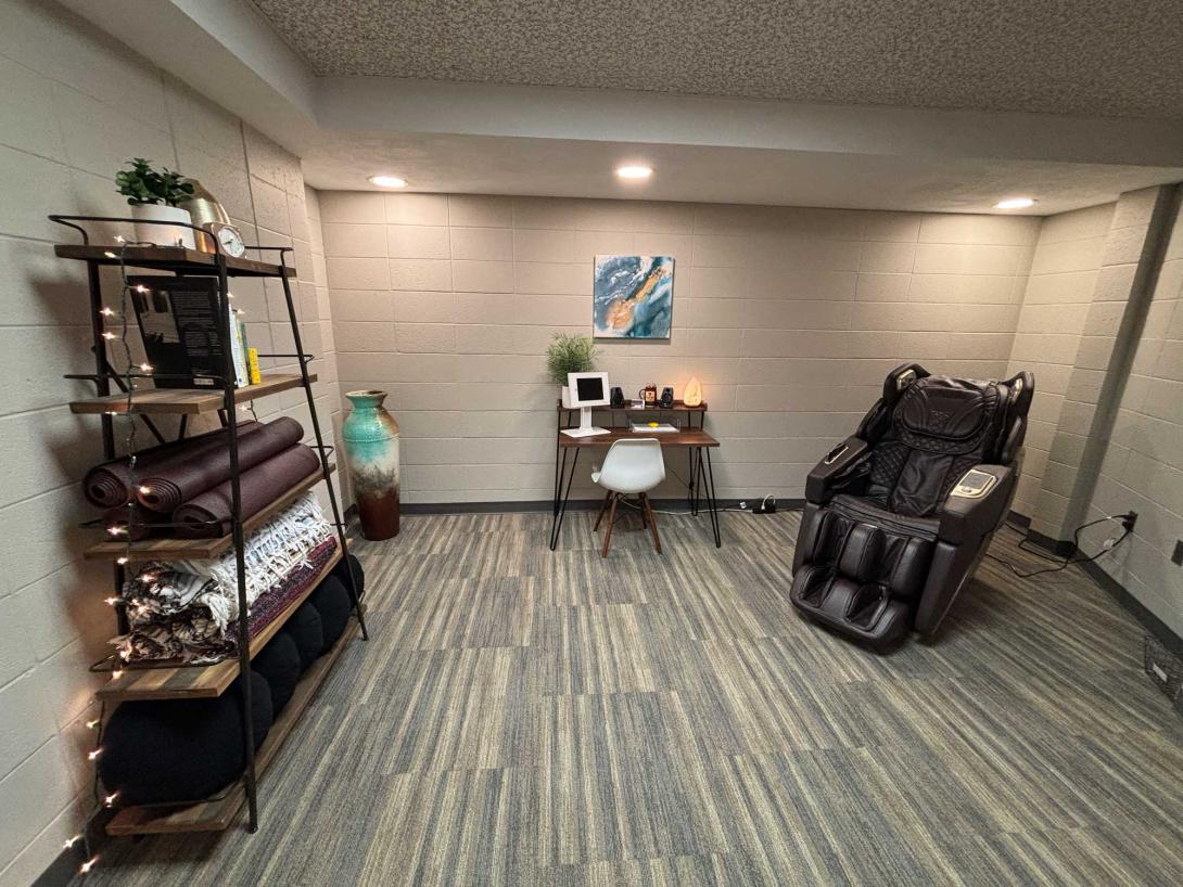 A room with a massage chair, desk with a salt lamp, and shelves with yoga mats and blankets.