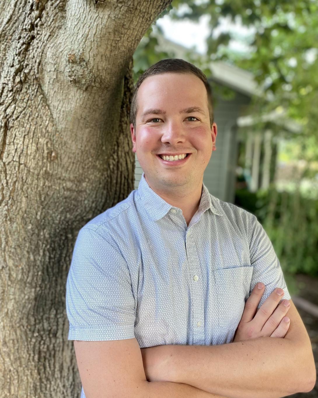 Caleb Lagerwey in a white short sleeved button down shirt leaning against a tree with his arms crossed