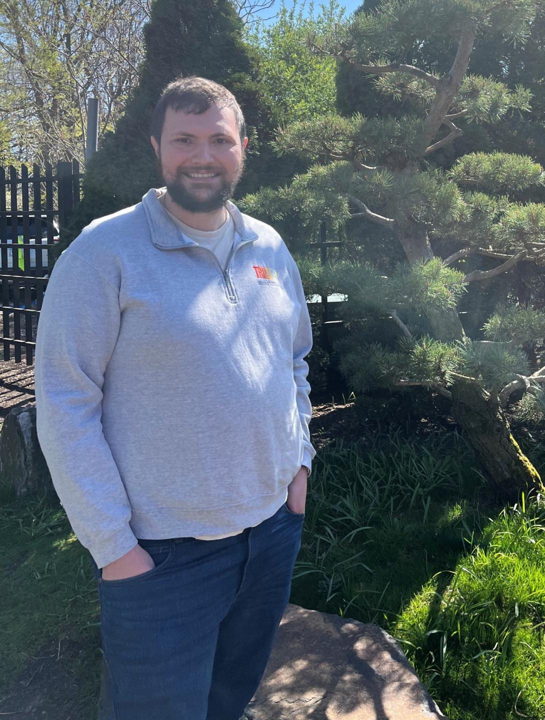Bryan Burke standing in a light gray pullover sweatshirt with his hands in his pockets in front of pine trees