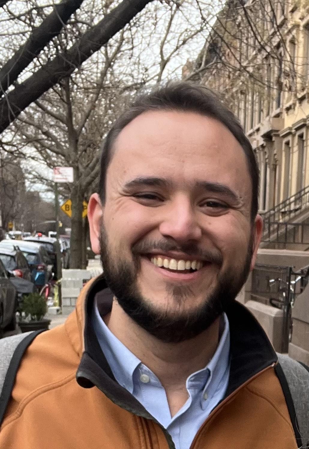 Close up photo of Isaac LaGrand smiling with leafless trees on a city street behind him