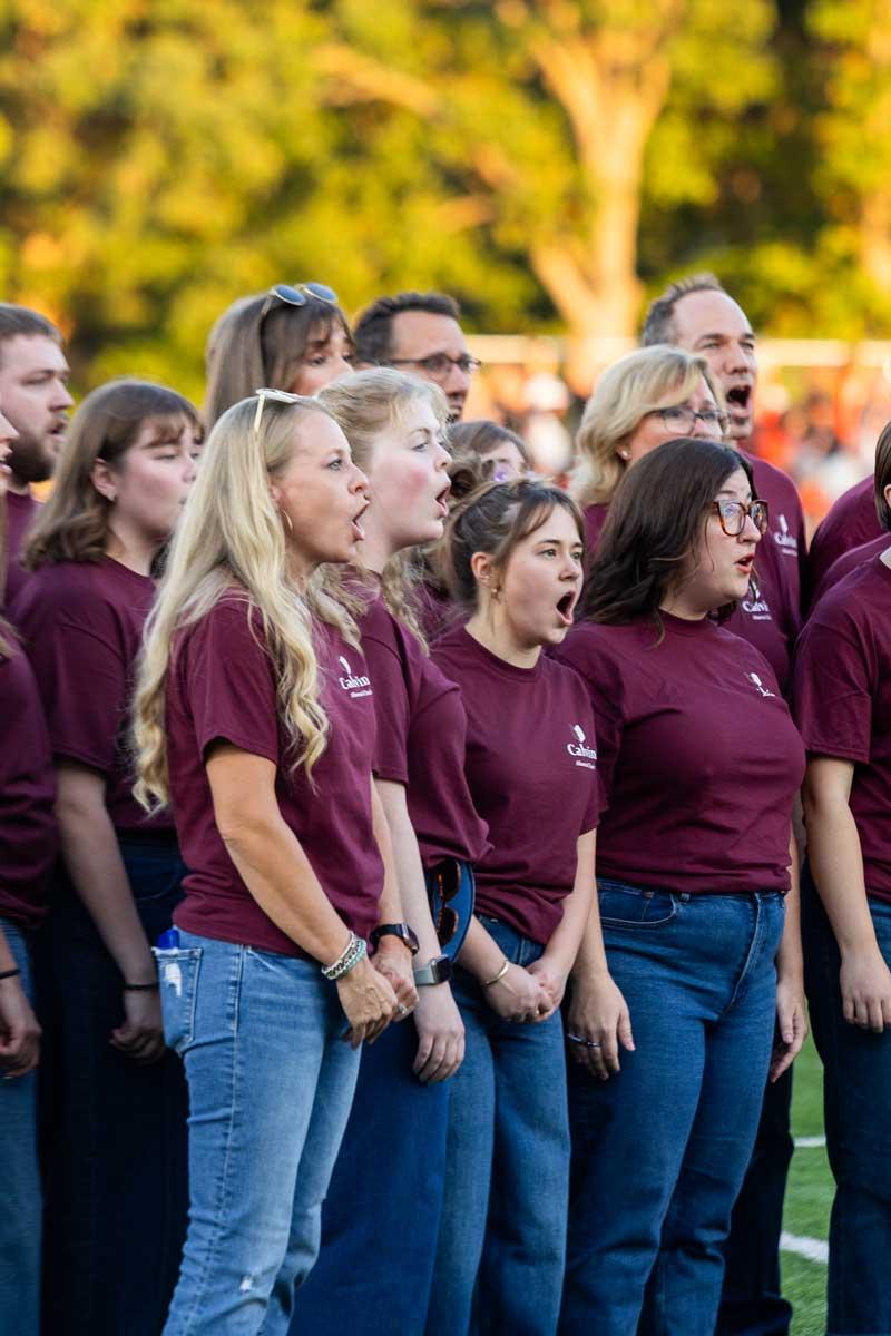 The Calvin Alumni Choir, wearing jeans and maroon shirts, sings the national anthem at the homecoming football game.