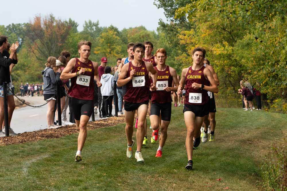 Calvin men's cross country runners race down a grass course, with spectators lining the street cheering them on.