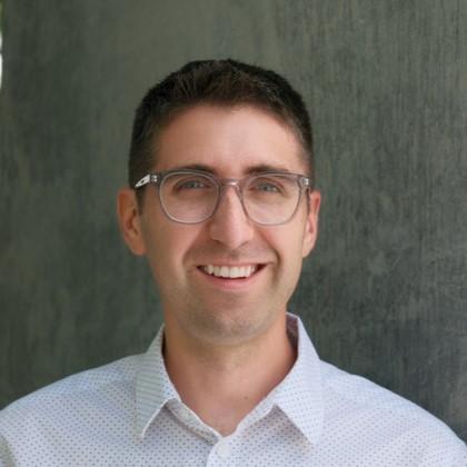 Dan Paulson in a white collared shirt with small black polka dots in front of a gray background
