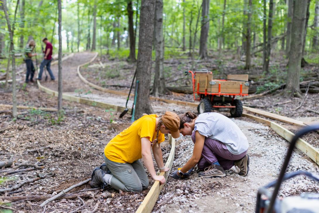 Two students kneel on a trail while drilling boards together to line the trail.