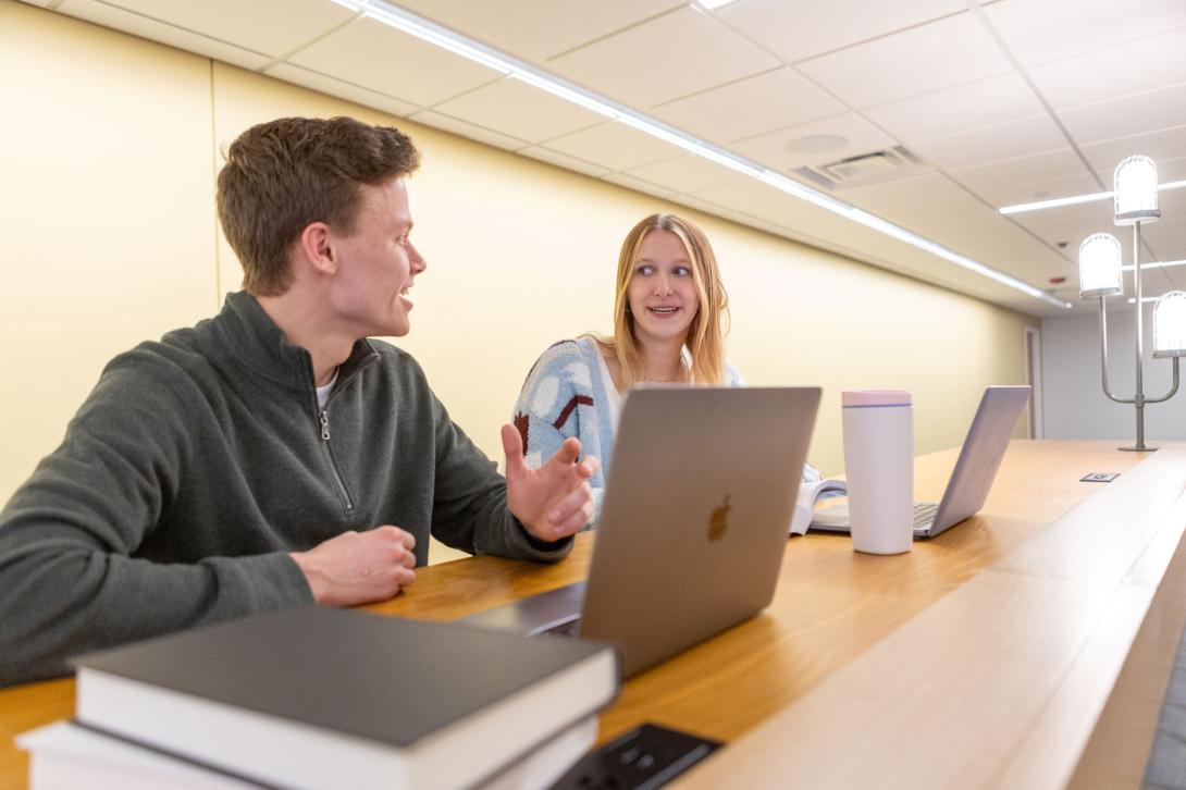 Two students sit at a table, having a conversation with laptops open.