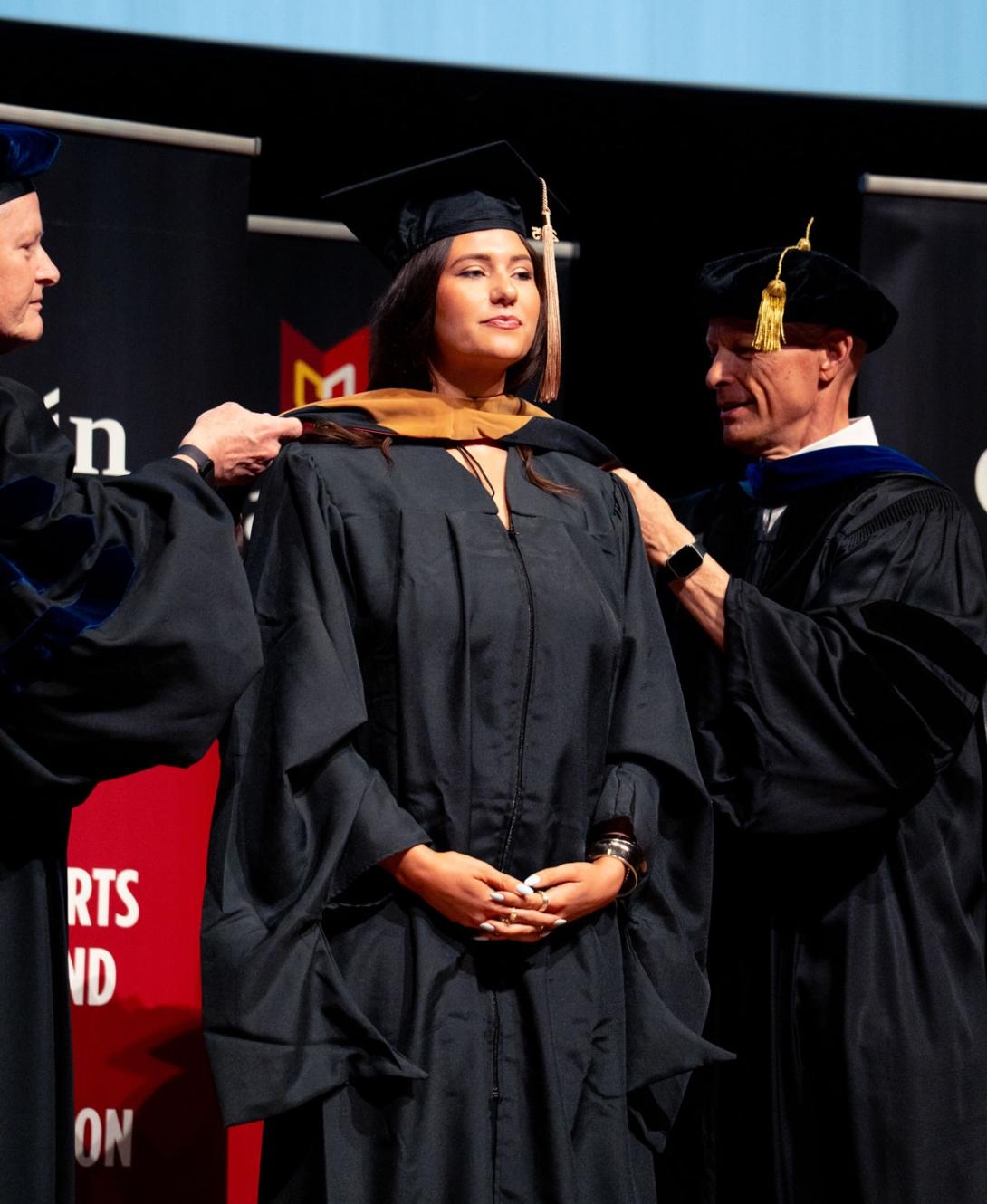 A female Calvin MBA student stands proudly on stage in cap and gown, with professors placing a master's hood over her shoulders.