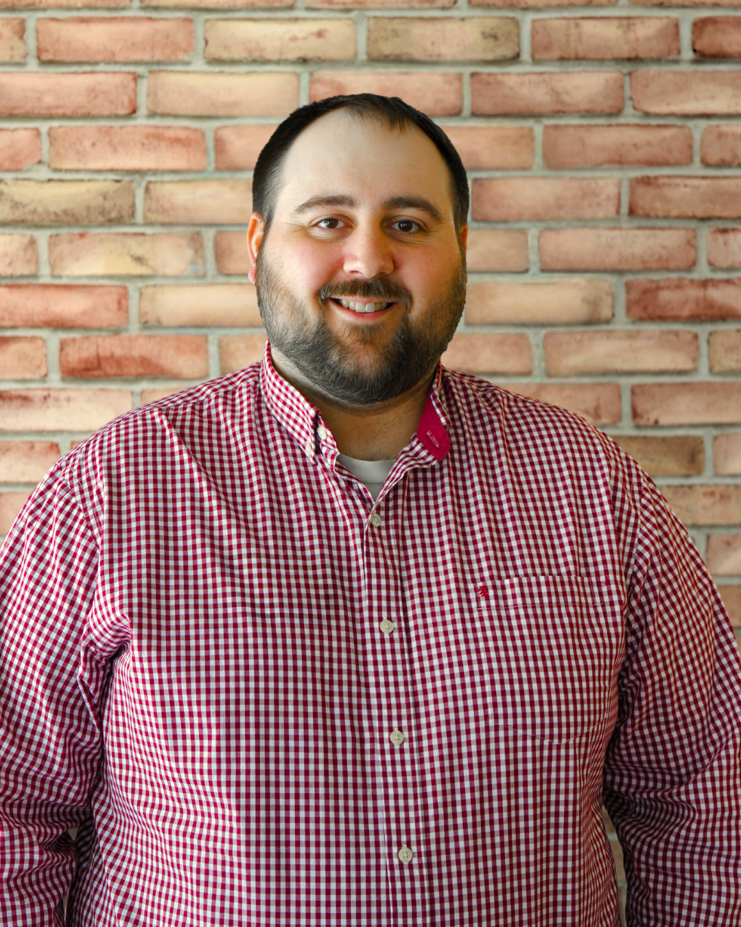 Man with dark hair and facial hair standing in front of a brick wall.