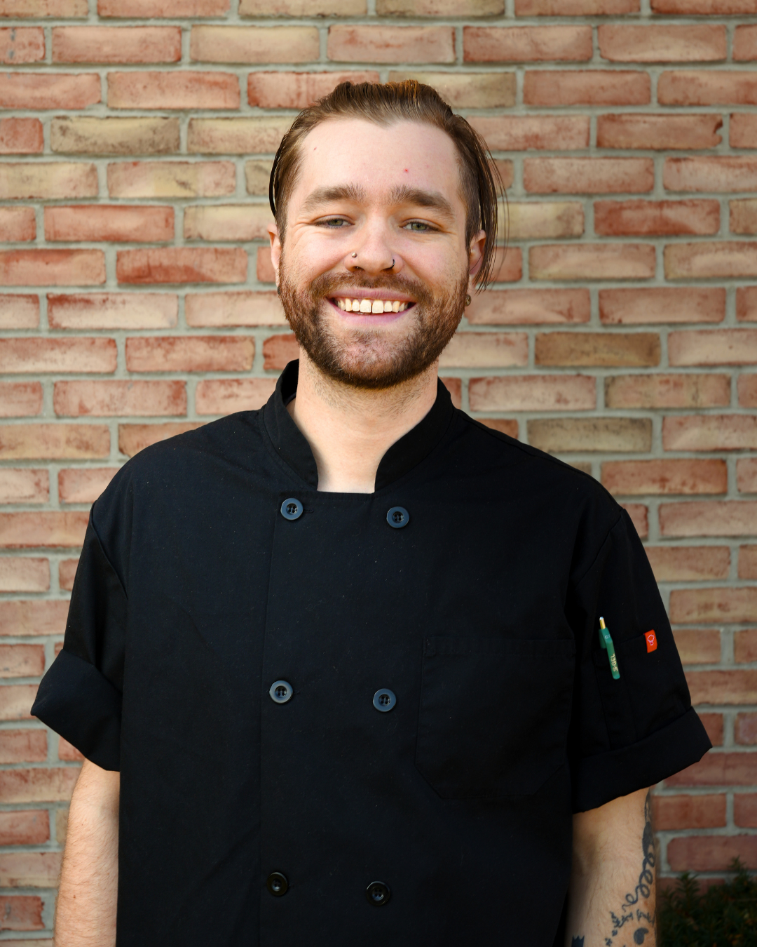 Man with facial hair wearing a black chef's coat, standing in front of a brick wall.