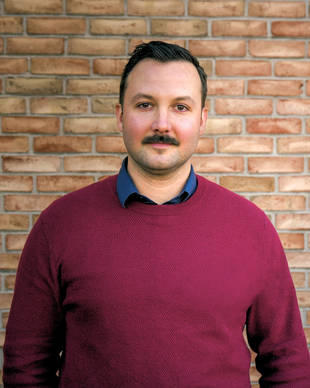 Man with dark hair and mustache wearing a burgundy sweater standing in front of a brick wall.