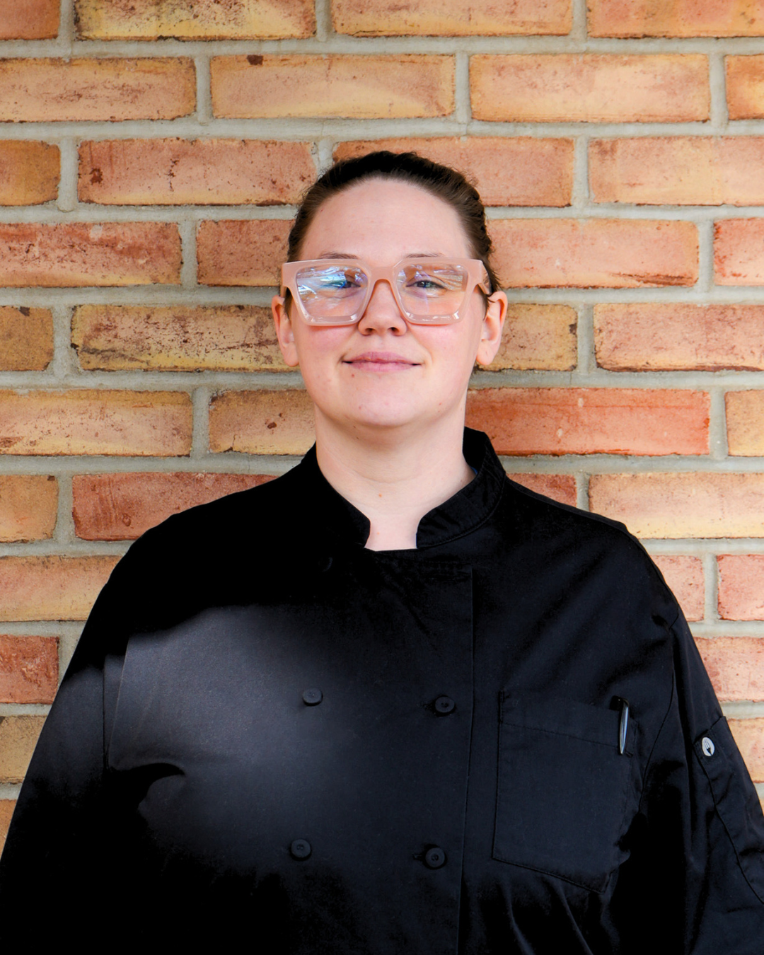 Woman with pink glasses wearing a chef's coat, standing in front of a brick wall.