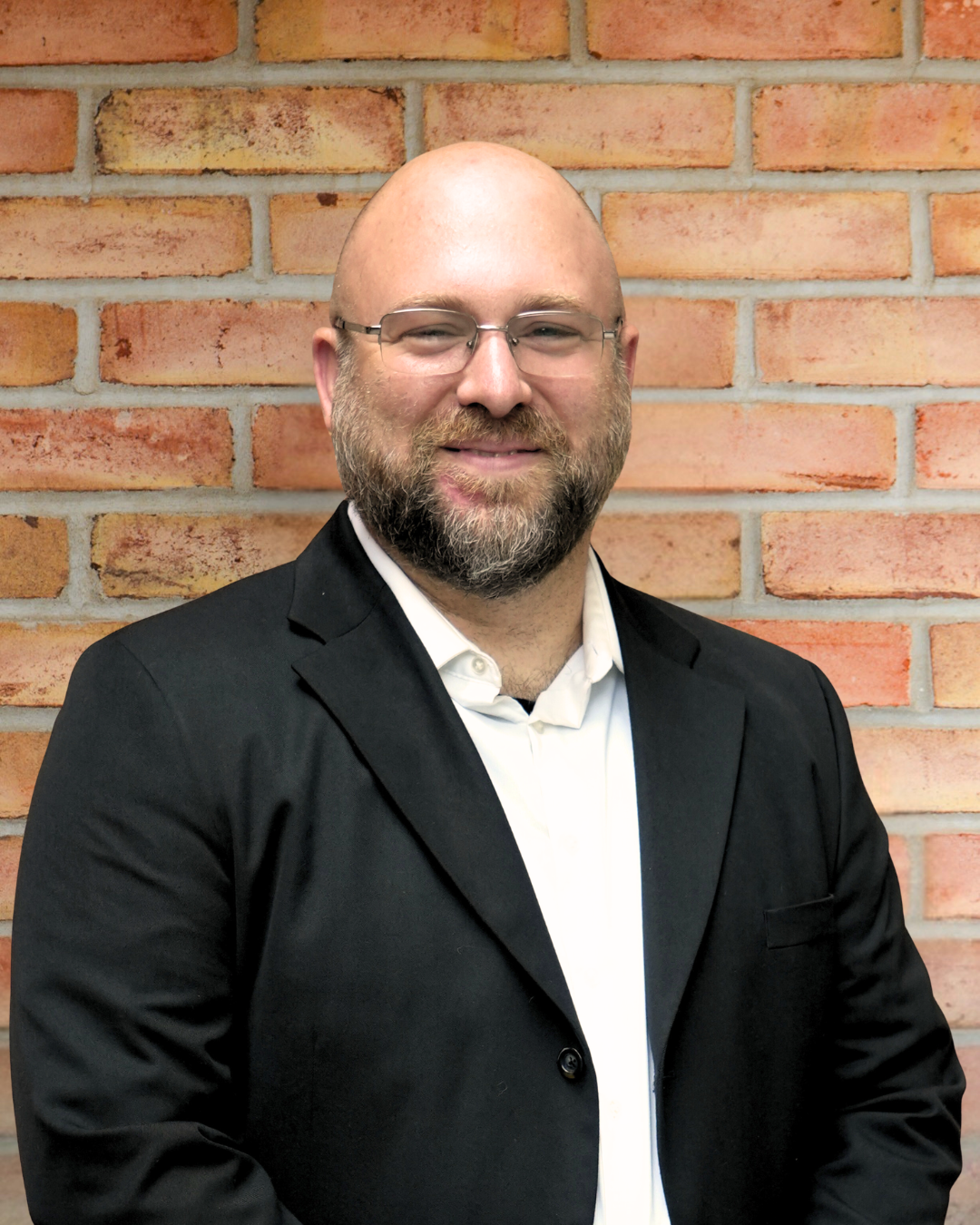Man with glasses and facial hair wearing a black jacket, standing in front of a brick wall.