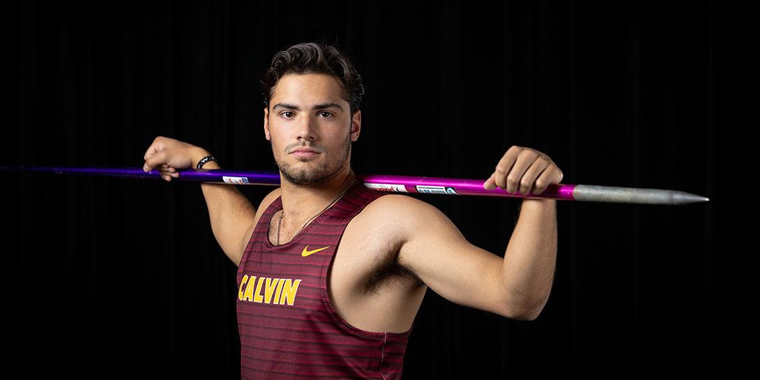Colin Jambekar poses with a serious, competitive face, wearing a Calvin track and field jersey and holding a javelin.