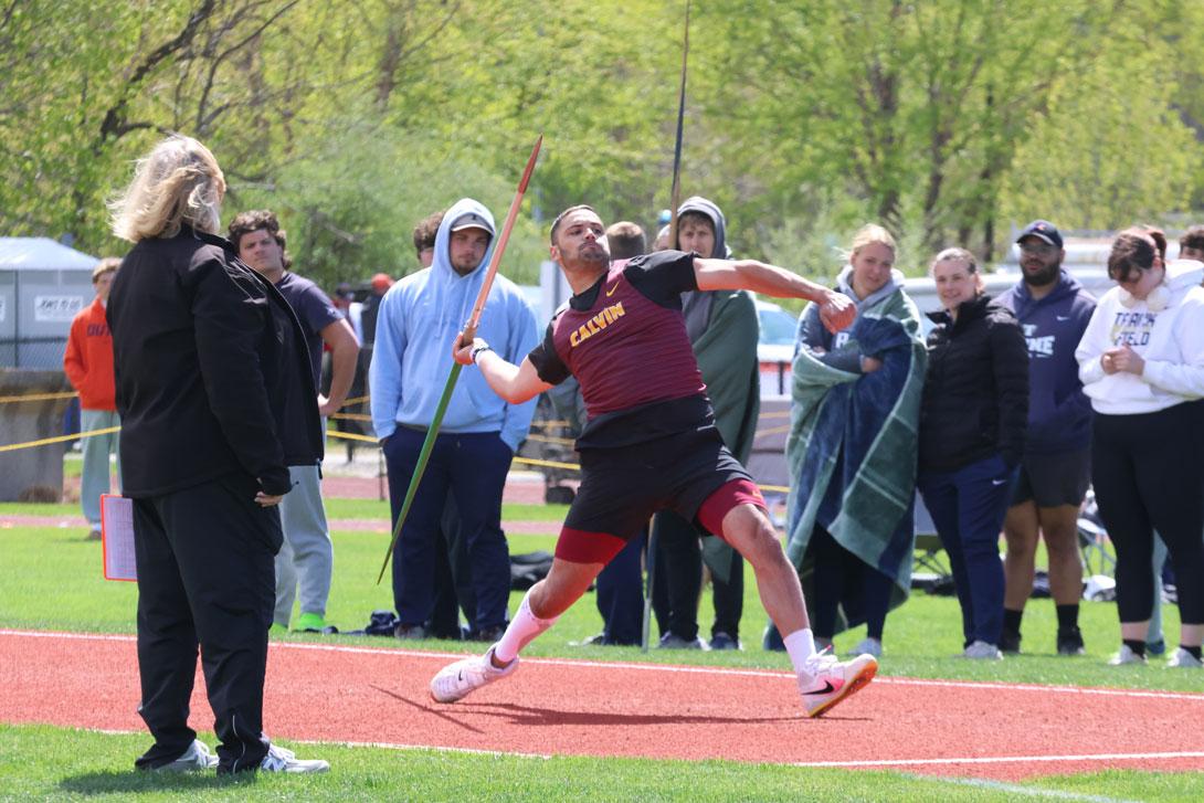 Colin Jambekar is about to release the javelin at a track and field competition, wearing a Calvin jersey, with fans cheering in the background.