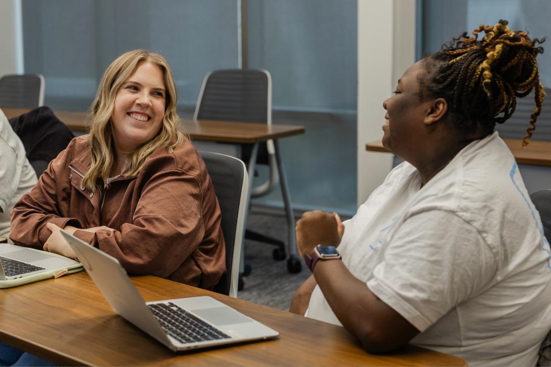 Two female students look at each other and smile. They are in a classroom with laptops open in front of each of them.