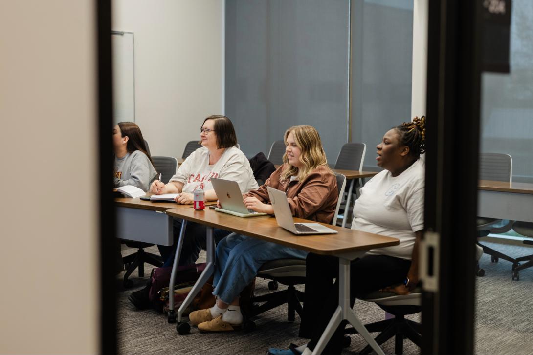 Four female students sit in a row at a long table in a college classroom, each with a laptop in front of them.