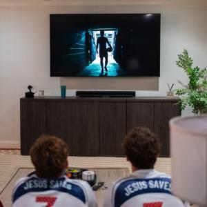 Two guys with Jesus Saves jerseys on watch a commercial on TV