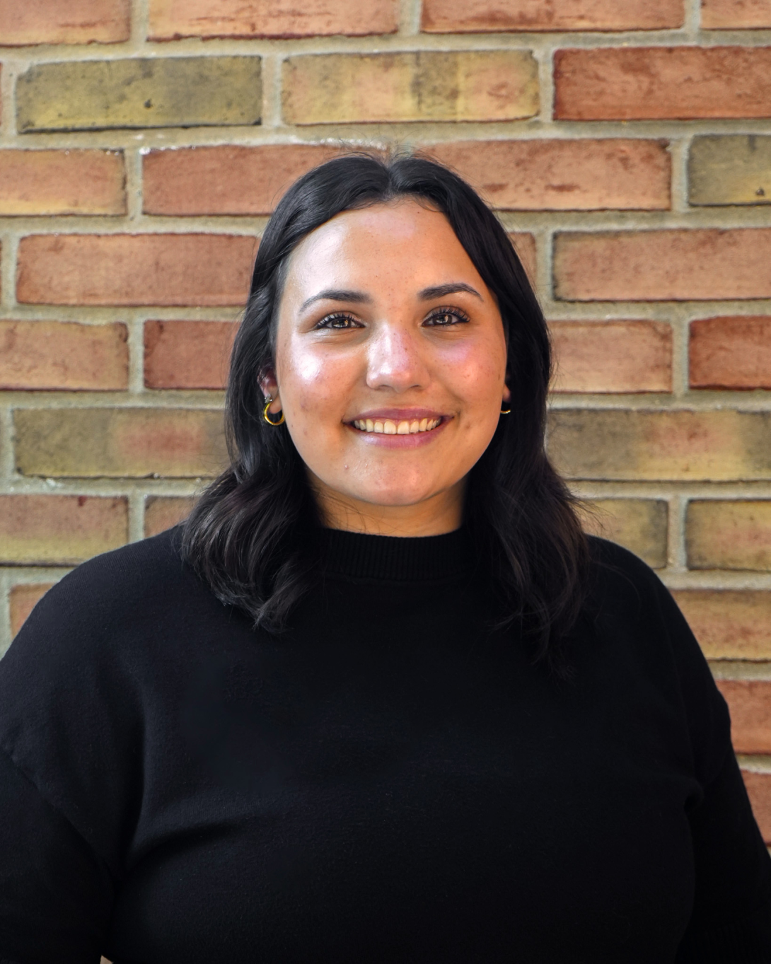 Woman in black shirt with brown hair smiling in front of a brick wall.
