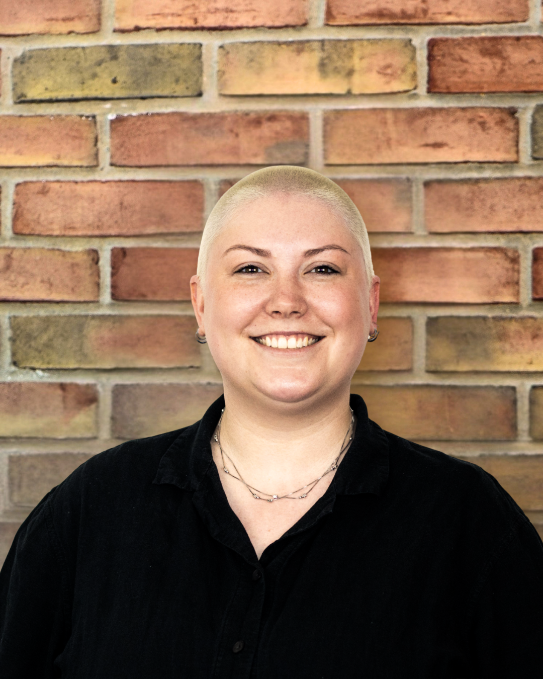 Woman in black shirt with blonde shaved head smiling in front of a brick wall.