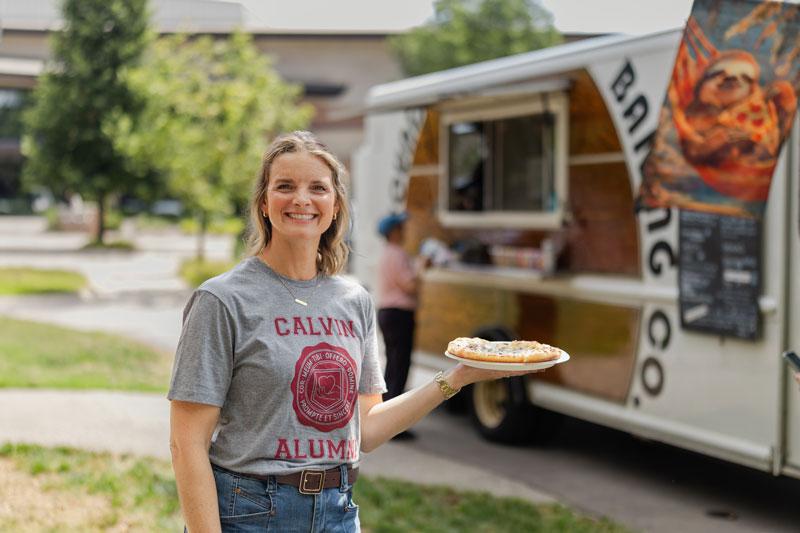 Becky Bagnall in a Calvin Alumni shirt smiles holding a pizza in front of a food truck on Calvin's campus.