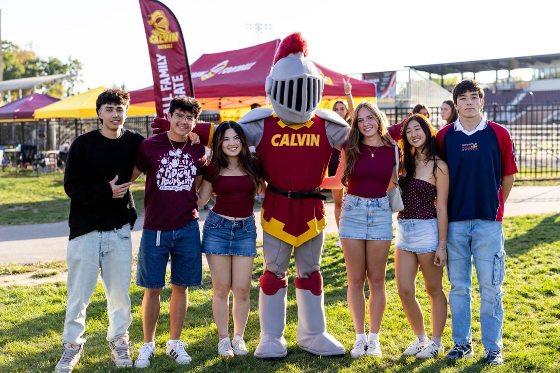 A group of students in maroon and gold stand with Joust the Calvin Knights mascot at a tailgate with tents in a field in the background.