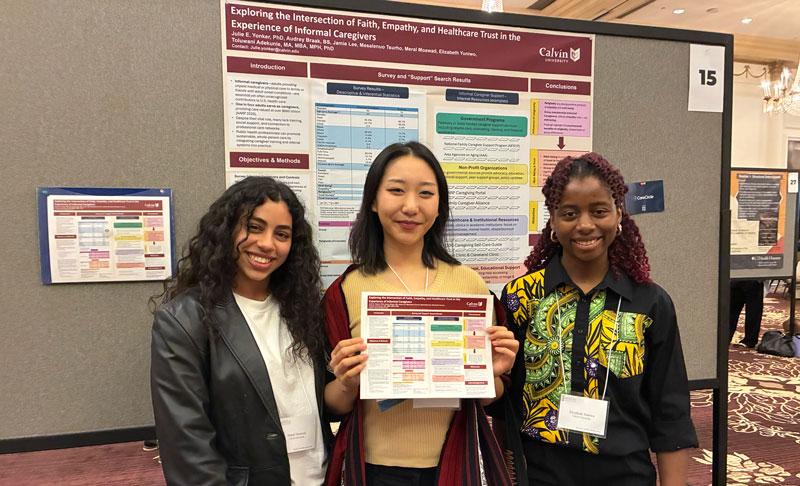 Three female student researchers stand in front of their research poster titled, Exploring the Intersection of Faith, Empathy, and Healthcare Trust in the Experience of Informal Caregivers.