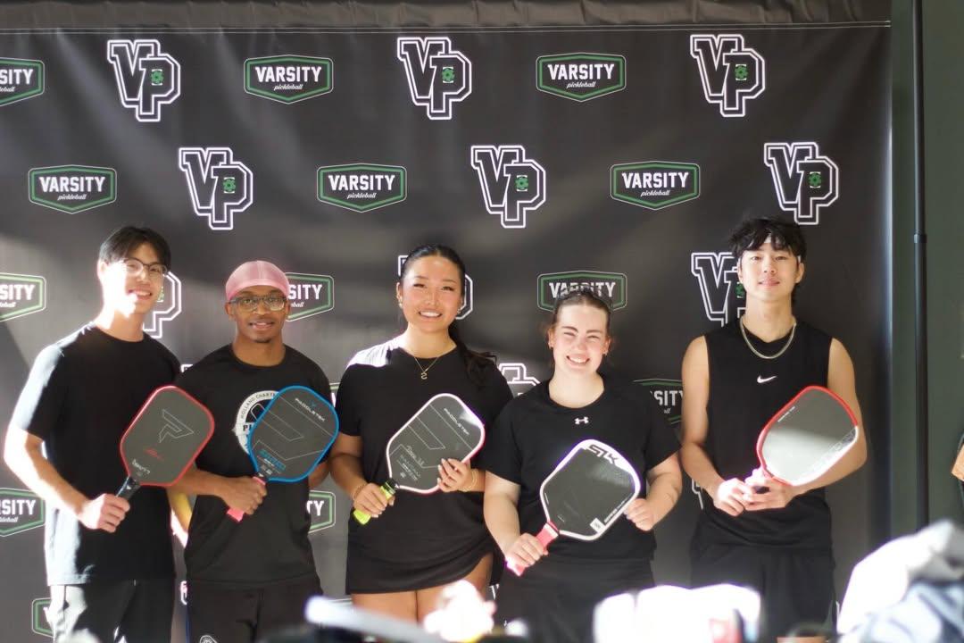 A group of five Calvin pickleball players dressed in black competition attire stand with paddles in front of a banner that says "varsity pickleball."
