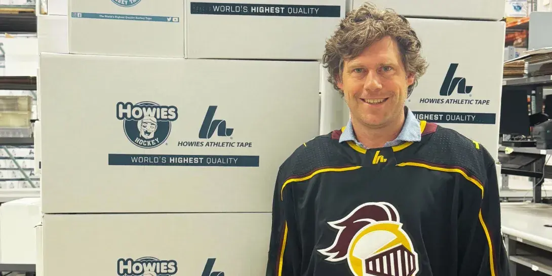 Max Sieplinga '04 standing with a Calvin hockey jersey on in front of Howie's HockeyTape boxes