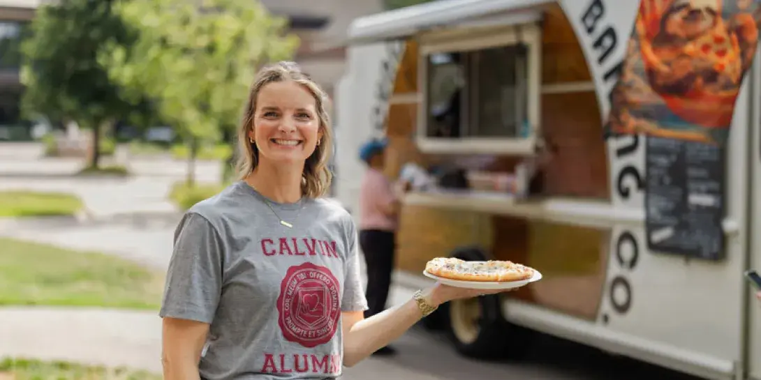 Becky Bagnall in a Calvin Alumni shirt smiles holding a pizza in front of a food truck on Calvin's campus.