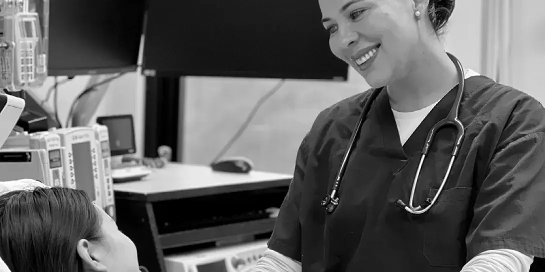 Laura Rivera in scrubs with a stethoscope stands and smiles at the bedside of a patient and her newborn.