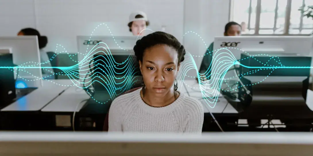 A young woman is focused on a computer in a lab with a few students seated behind her at computers.