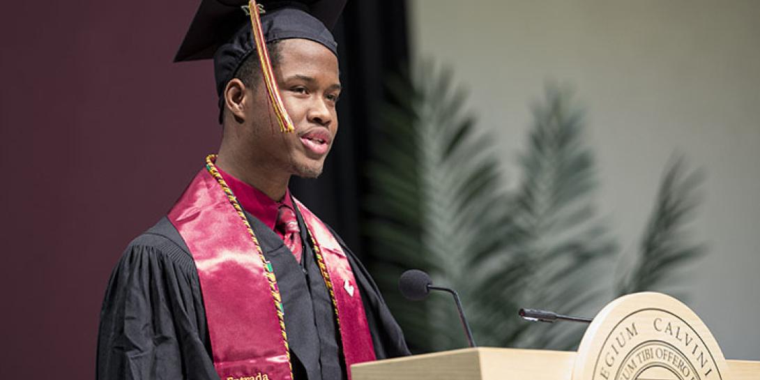 Student in cap and gown standing at a podium provides remarks at formal Commencement ceremony.