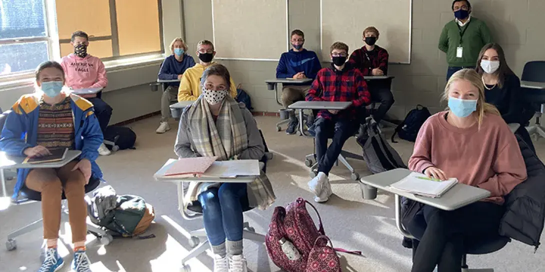 Class of high school students in masks sit in desks with college professor standing in back of room