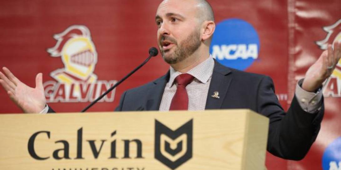 Head Football Coach Trent Figg speaking behind Calvin Univerity podium in suit and tie.