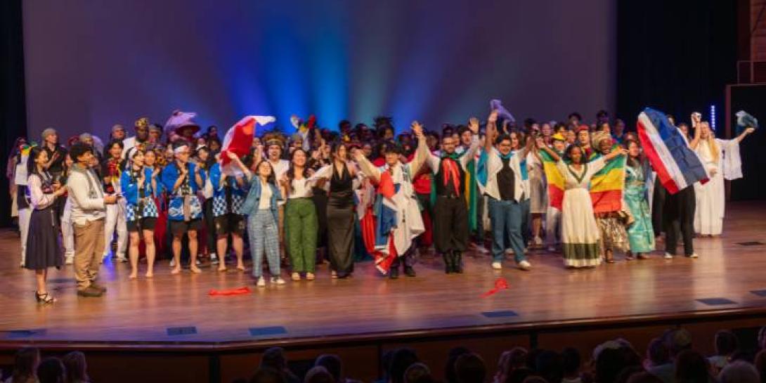 A large group of students holding flags stands on stage after a performance.